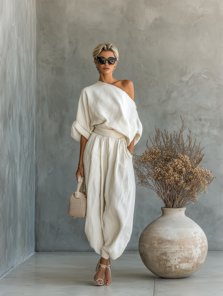 Woman in elegant, flowing off-shoulder white outfit with a beige woven bag, standing next to a large ceramic vase with dried flowers indoors.