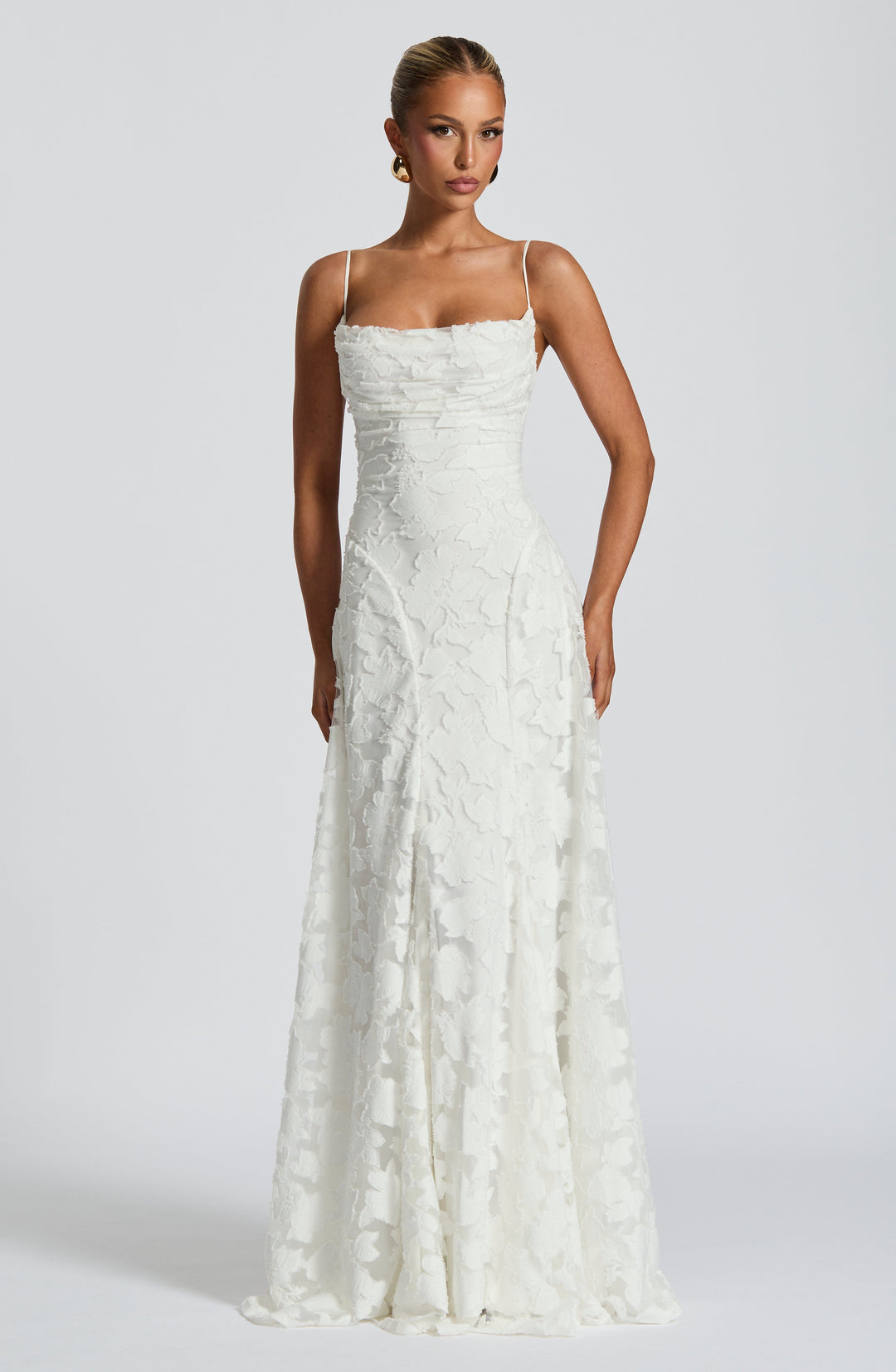 Elegant maxi dress in white with floral texture, thin straps, and floor-length design in a studio setting.