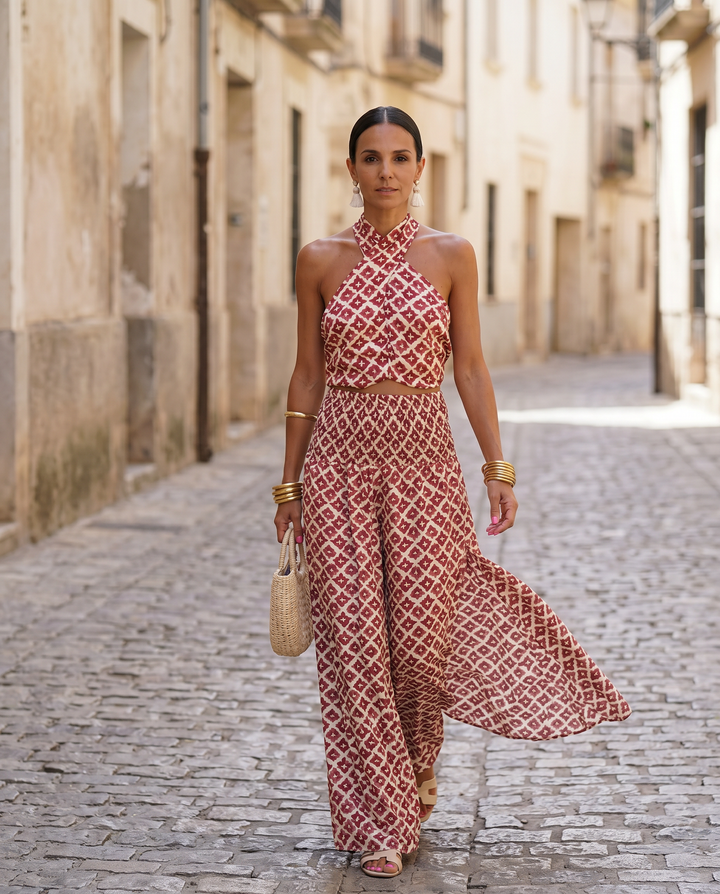 Woman wearing patterned red and white halter top and matching wide-leg pants, holding woven bag, walking on cobblestone street.