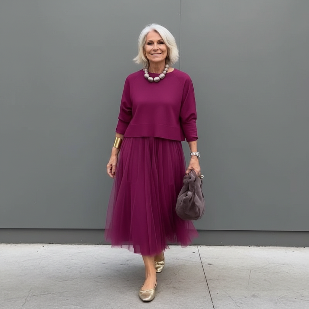 Stylish woman in burgundy pleated skirt outfit with beads, standing by a gray wall.