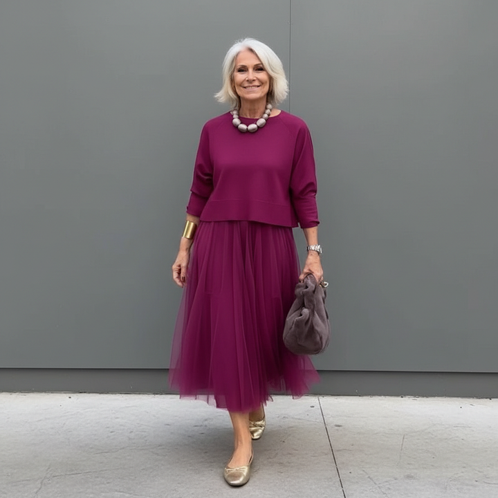 Stylish woman in burgundy pleated skirt outfit with beads, standing by a gray wall.