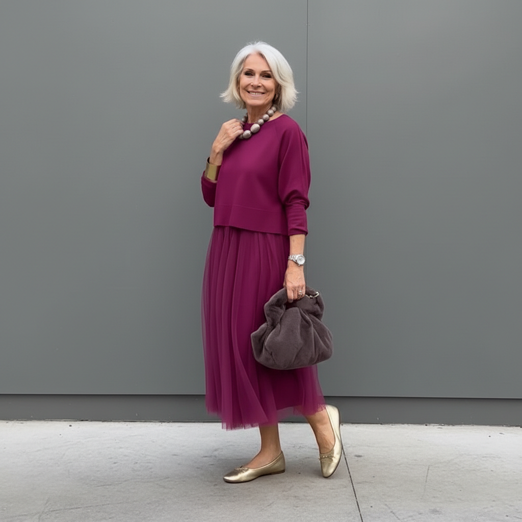 Elegant woman in burgundy outfit with pleated skirt and beaded necklace, outdoor background.
