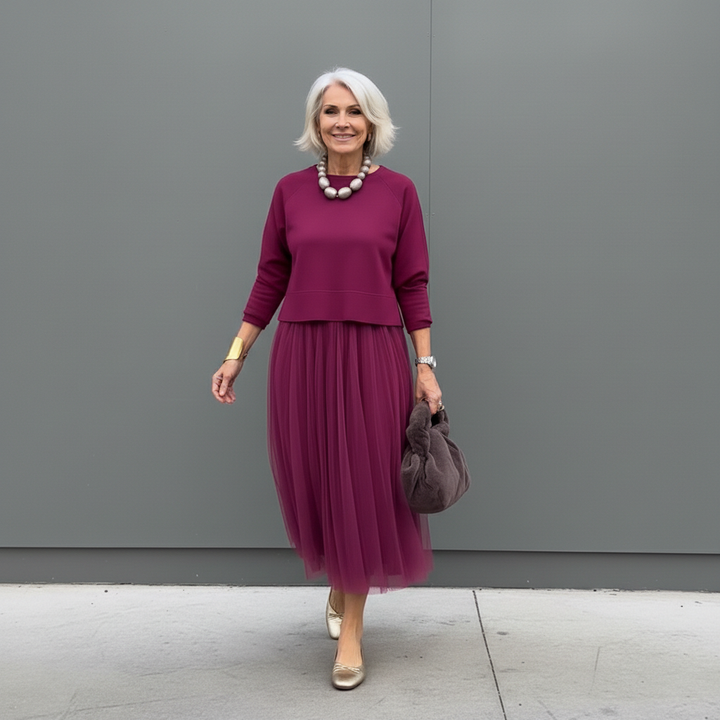 Elegant woman in burgundy two-piece set with pleated skirt, accessorized with beads, outdoor setting.