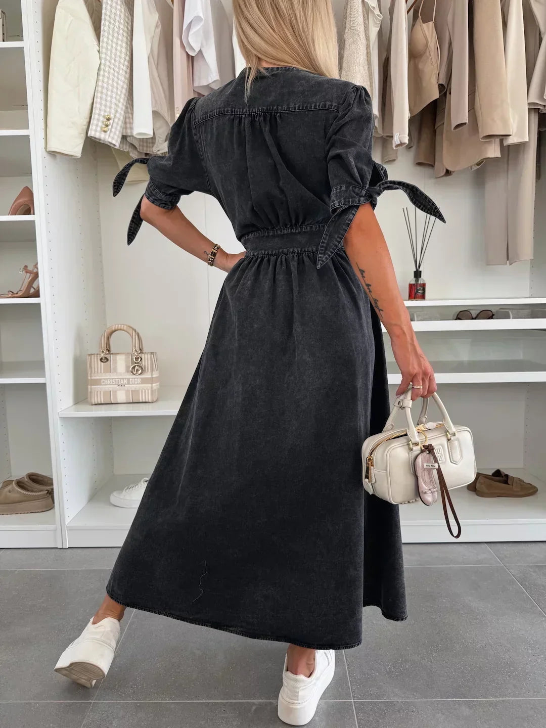 Back view of black denim dress with tied sleeves, worn by a person holding a white bag, in a closet setting.