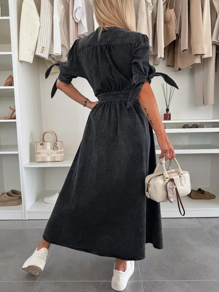 Back view of black denim dress with tied sleeves, worn by a person holding a white bag, in a closet setting.