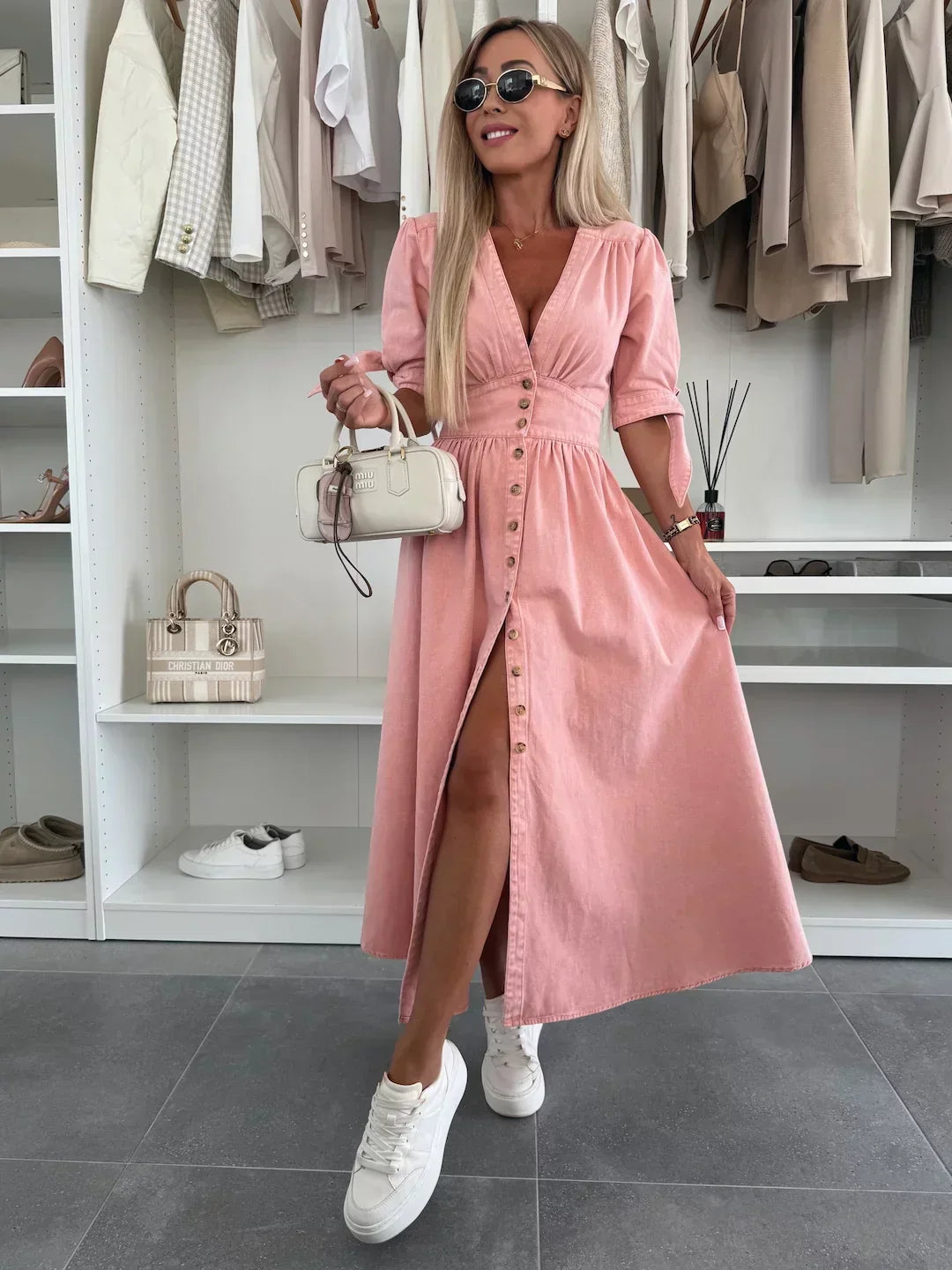 Pink denim dress with buttons and tied sleeves, worn by a person with sunglasses and white sneakers, in a closet setting.
