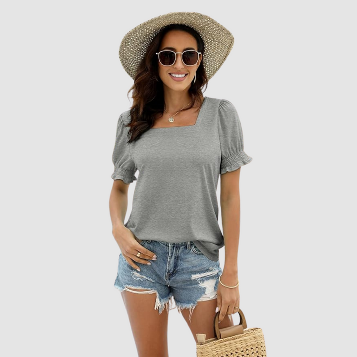 Woman in gray square neck top, ripped denim shorts, straw hat, sunglasses, holds woven bag, neutral backdrop.