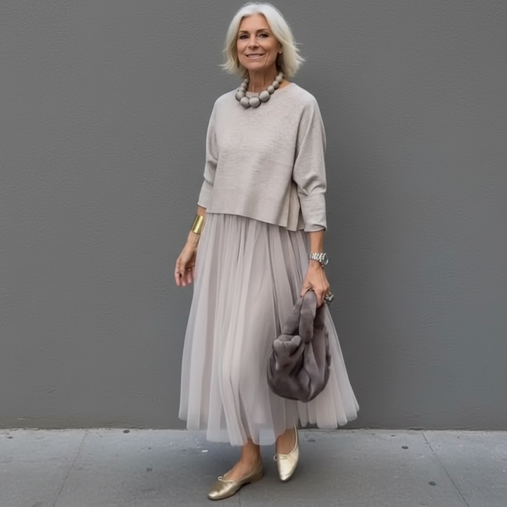 Sophisticated woman in gray pleated skirt outfit, with a plush bag, standing in an urban area.