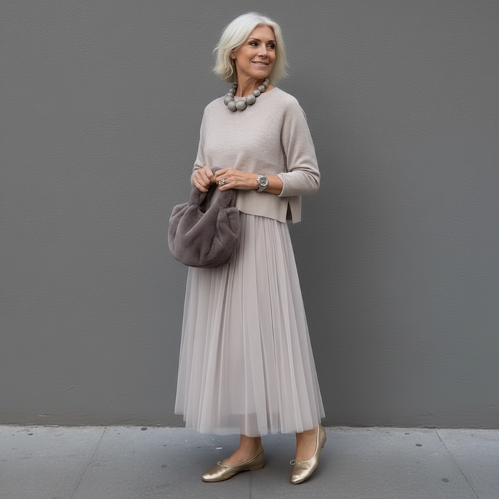 Elegant woman in gray outfit with pleated skirt and a plush handbag, standing outdoors.