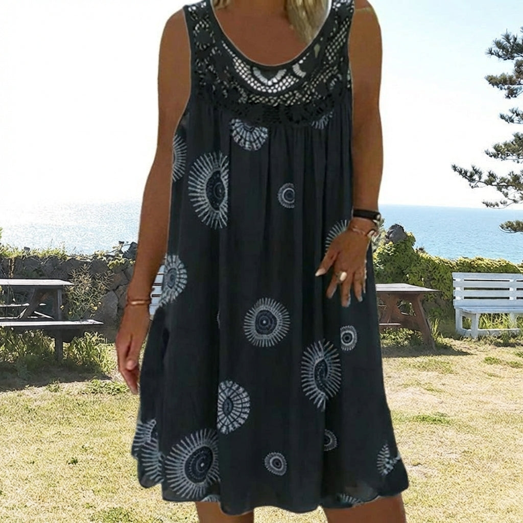 Woman in black printed dress with ocean view and benches outdoors.