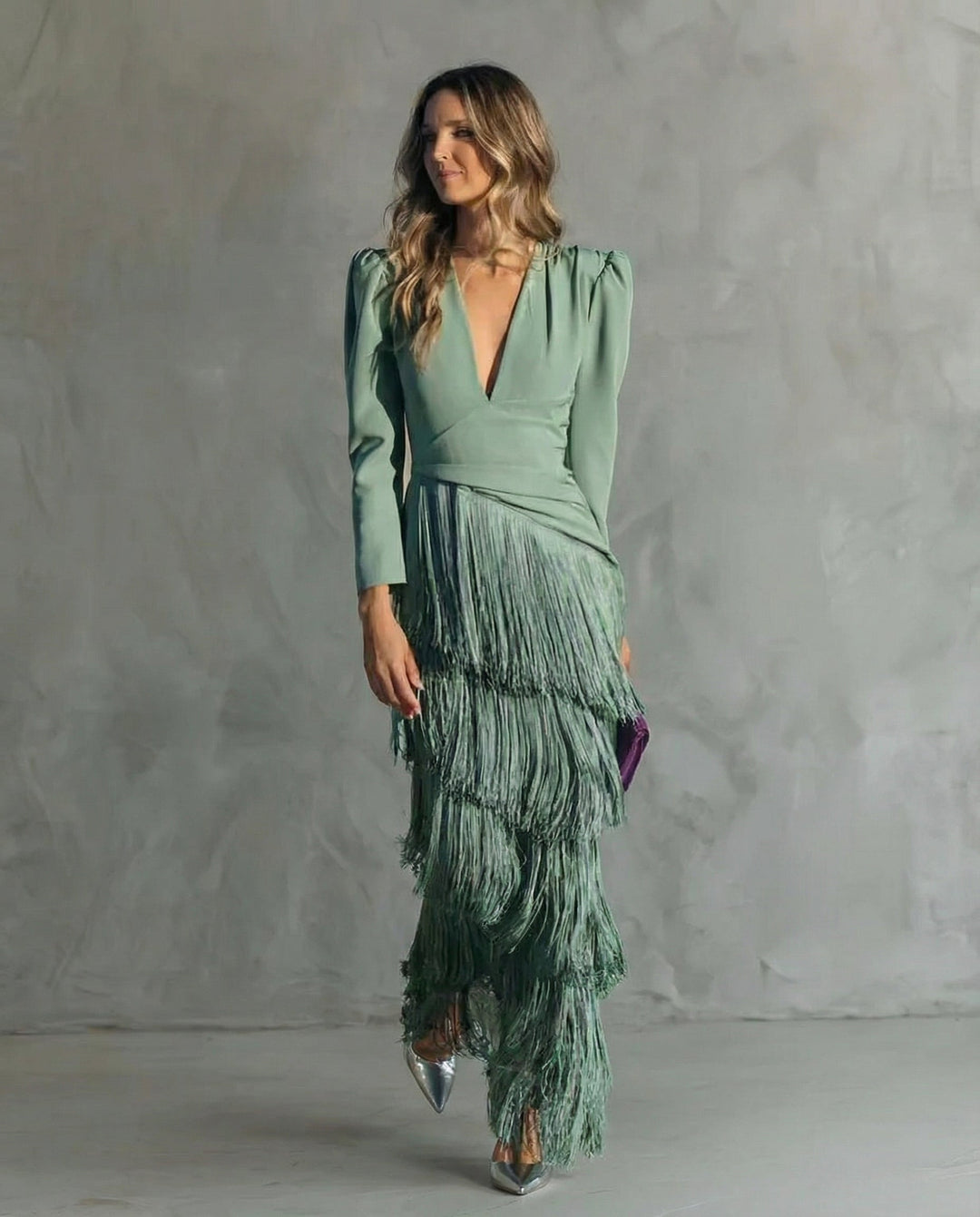 A woman in a sage green dress with a fringed skirt, standing in a minimalist room with a textured gray wall.