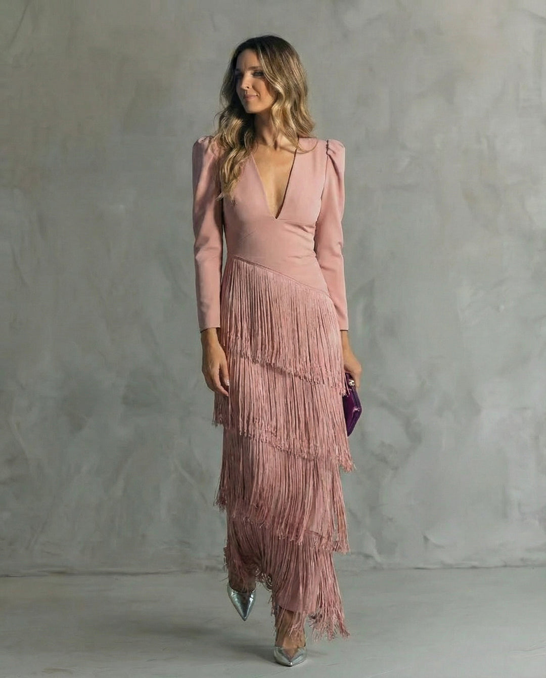 A woman in a blush pink dress with layered fringe, holding a purple clutch, standing against a gray textured background.