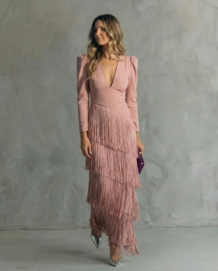 A woman in a blush pink dress with layered fringe, holding a purple clutch, standing against a gray textured background.