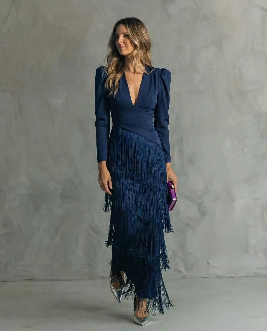 A woman in a navy blue dress with fringe details, holding a purple clutch, standing in a minimalist room with gray walls.