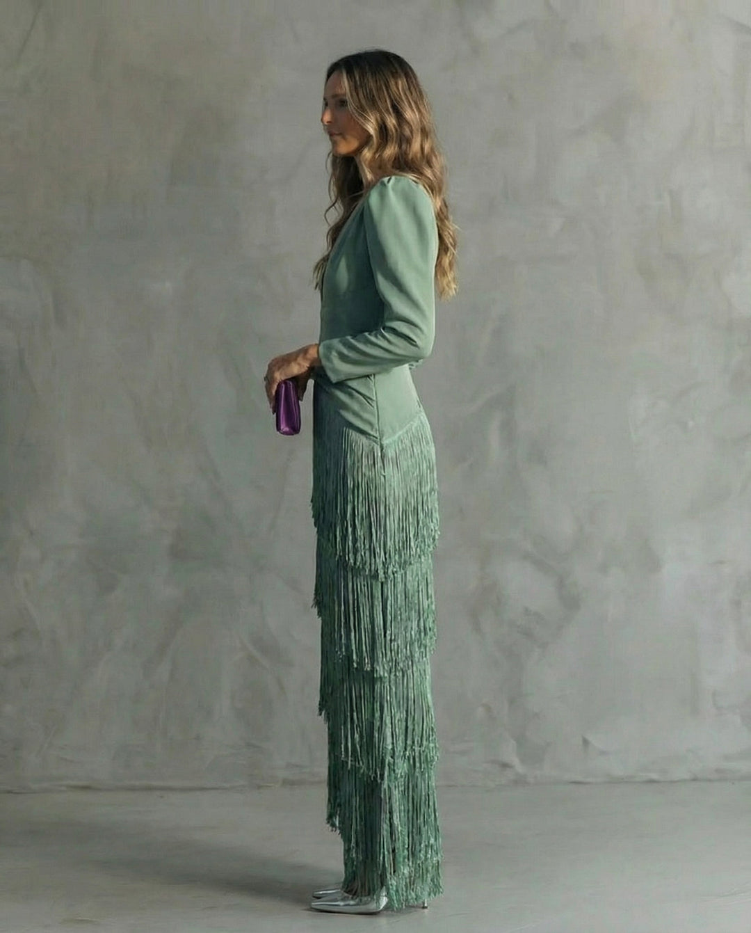 Side view of a woman in a sage green dress with fringe details, holding a purple clutch against a gray backdrop.