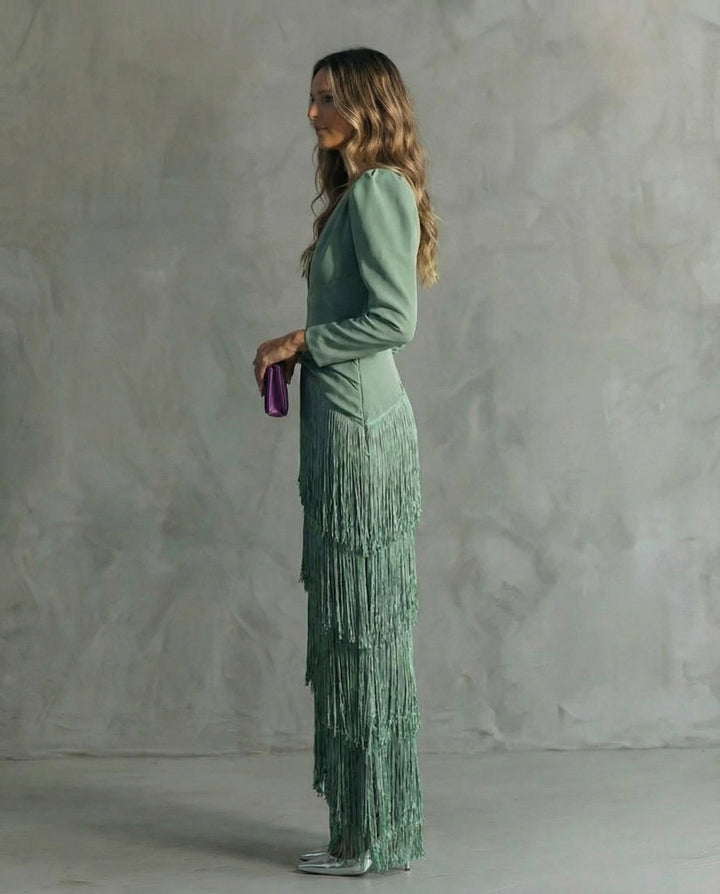 Side view of a woman in a sage green dress with fringe details, holding a purple clutch against a gray backdrop.