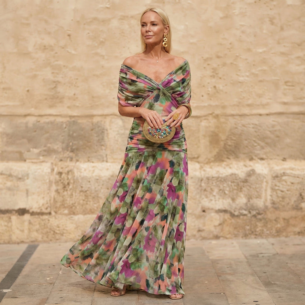 Woman wearing an off-shoulder, flowing floral dress, holding a decorated clutch, standing against a textured stone wall.