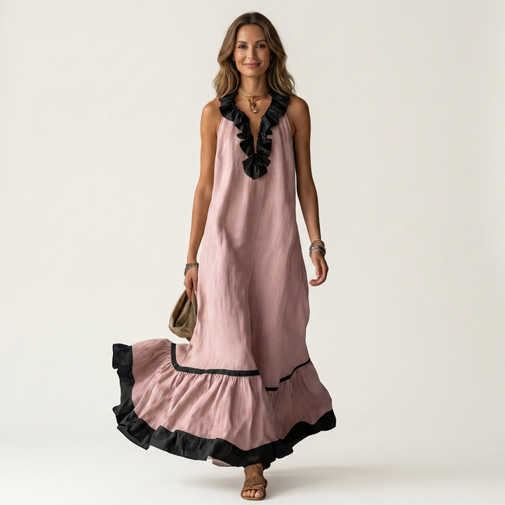 Woman in pink sleeveless maxi dress with ruffle-neck and contrast trim, holding bag, smiling in studio.