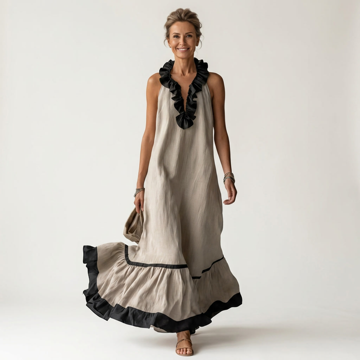 Woman in beige sleeveless maxi dress with ruffle-neck and contrast trim, holding bag, smiling in studio.