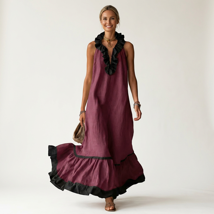 Woman in burgundy sleeveless maxi dress with ruffle-neck and contrast trim, holding bag, smiling in studio.