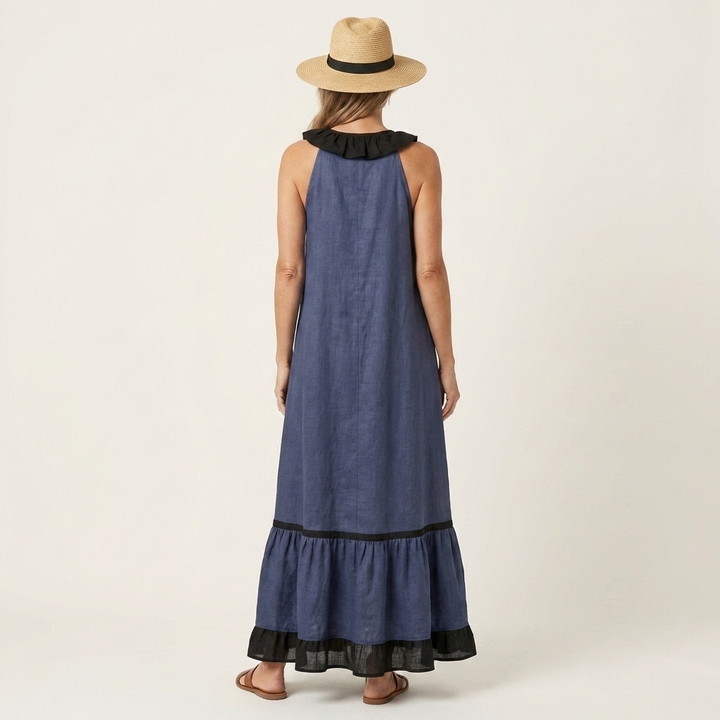 Back view of woman in dark blue sleeveless maxi dress with ruffle-neck and contrast trim, wearing hat in studio.