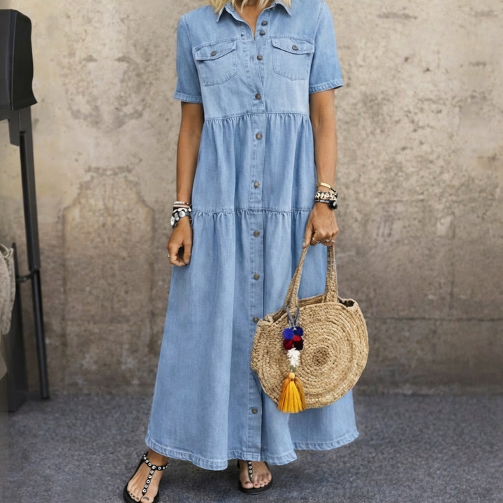 Light blue button-front midi dress with short sleeves, tiered design, and front pockets. Worn with a round straw bag in a textured studio setting.
