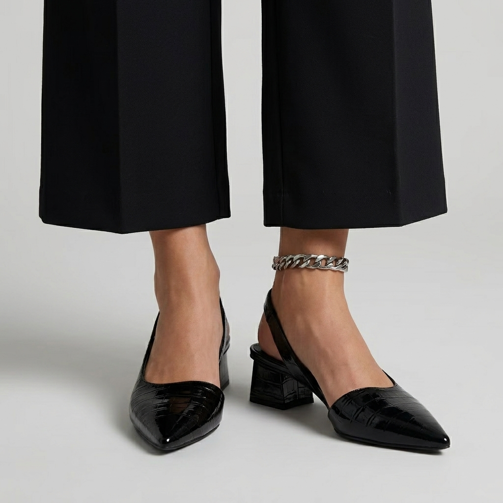 Person wearing black orthopedic heels with a shiny, textured finish and cropped black trousers in a studio setting.