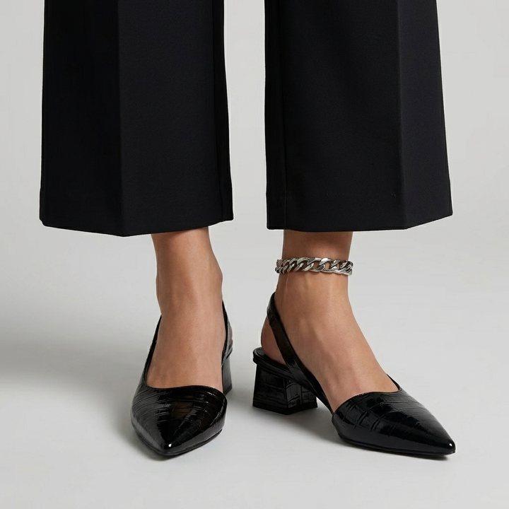 Person wearing black orthopedic heels with a shiny, textured finish and cropped black trousers in a studio setting.