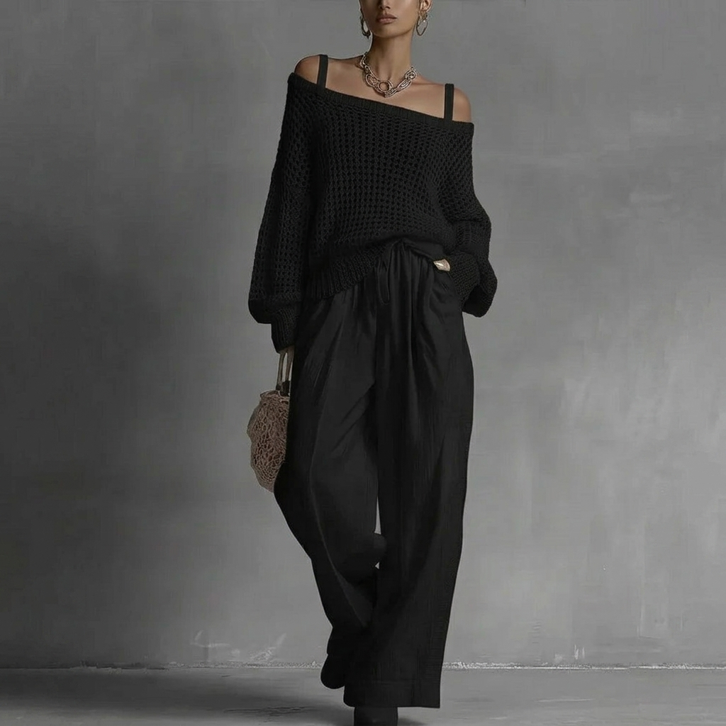Woman in off-shoulder black top and wide black pants, holding woven bag in minimal gray studio.