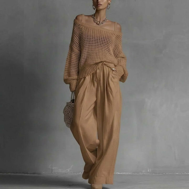Woman in off-shoulder tan top and wide tan pants, holding woven bag in minimal gray studio.