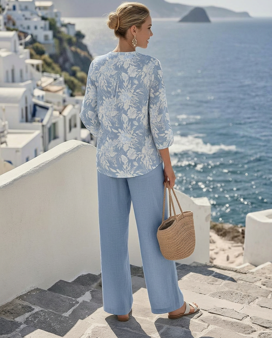Back view of a woman in a blue floral outfit with a straw bag, standing on stairs with ocean backdrop.