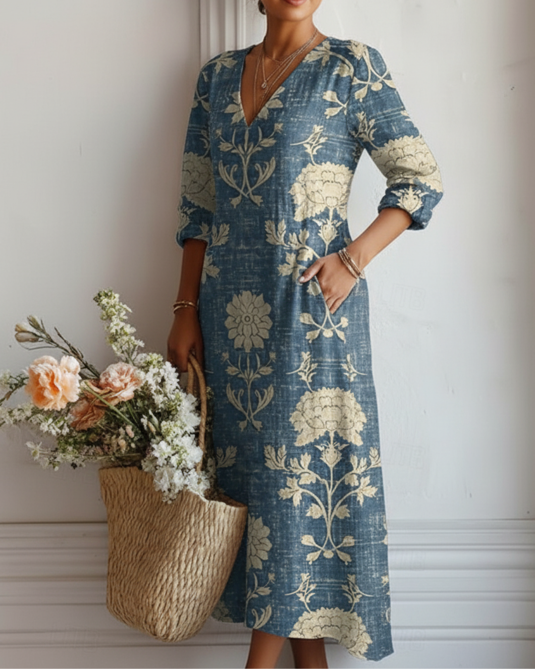 Blue dress with floral design worn by woman, posing with hand on hip, holding basket of flowers, in white room.