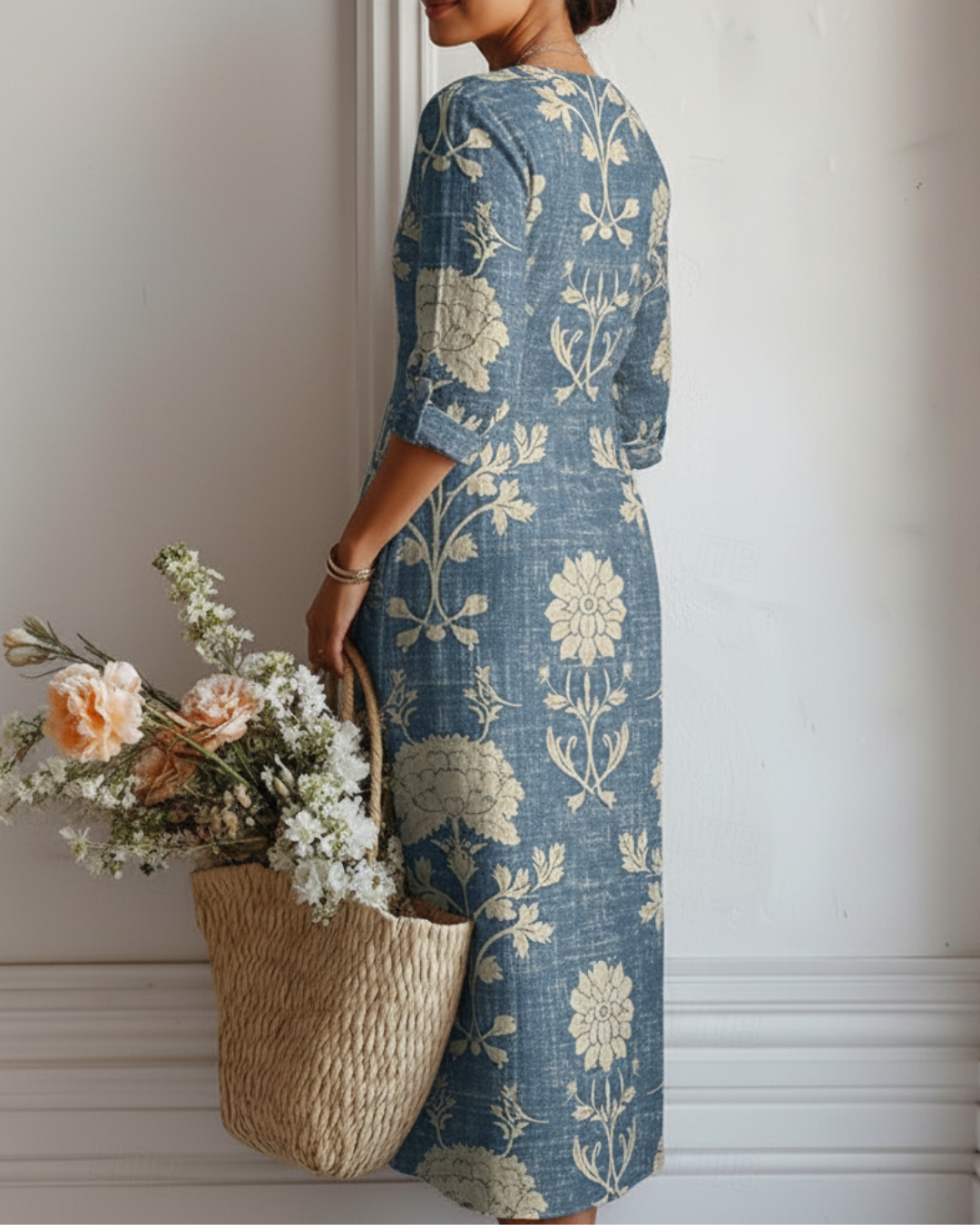 Back view of woman in floral blue dress, holding a basket of flowers, standing against white wall indoors.
