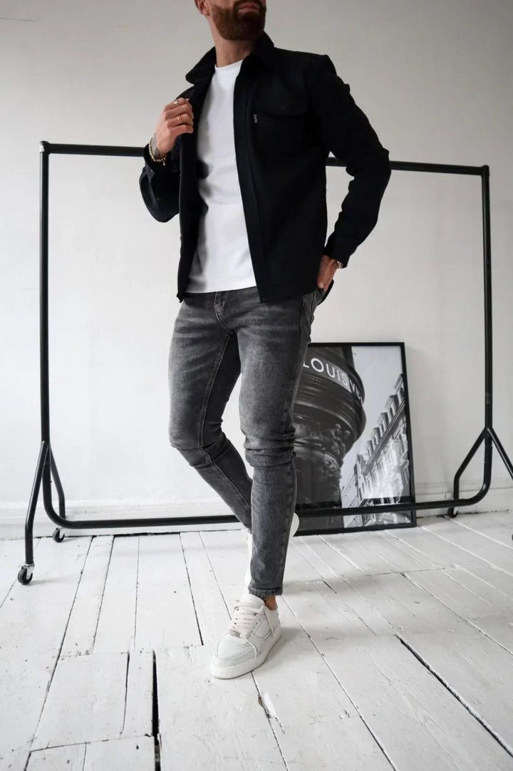 Black overshirt over a white t-shirt and jeans, standing indoors beside rail and artwork.