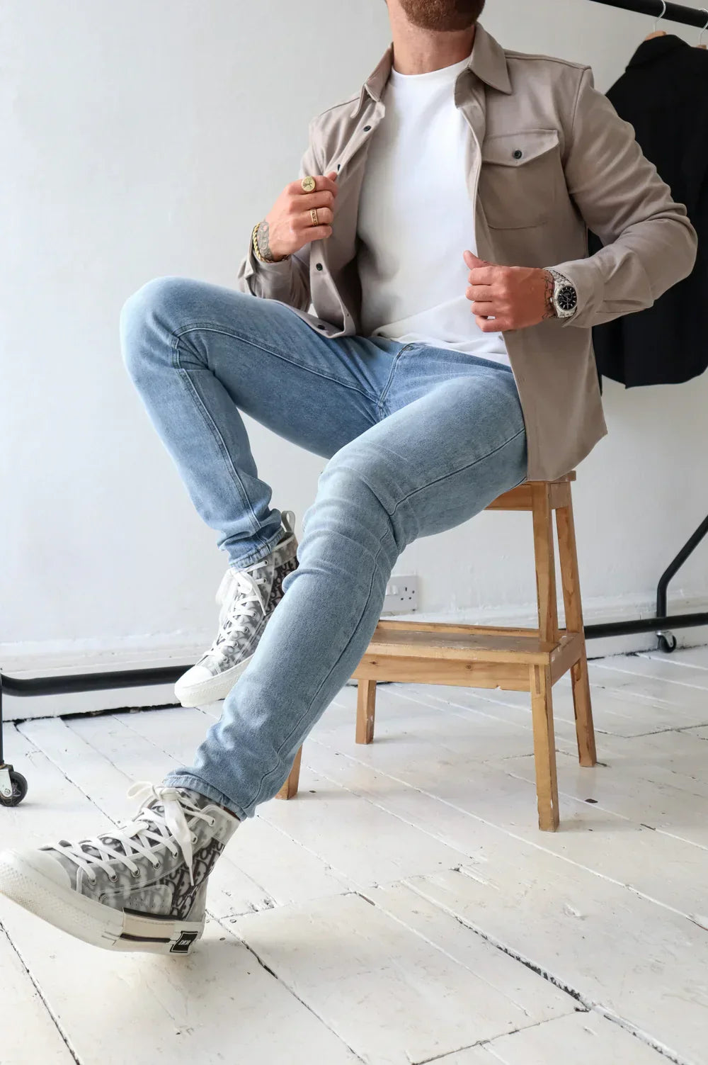 Taupe overshirt with white t-shirt and light jeans, seated on wooden stool indoors.
