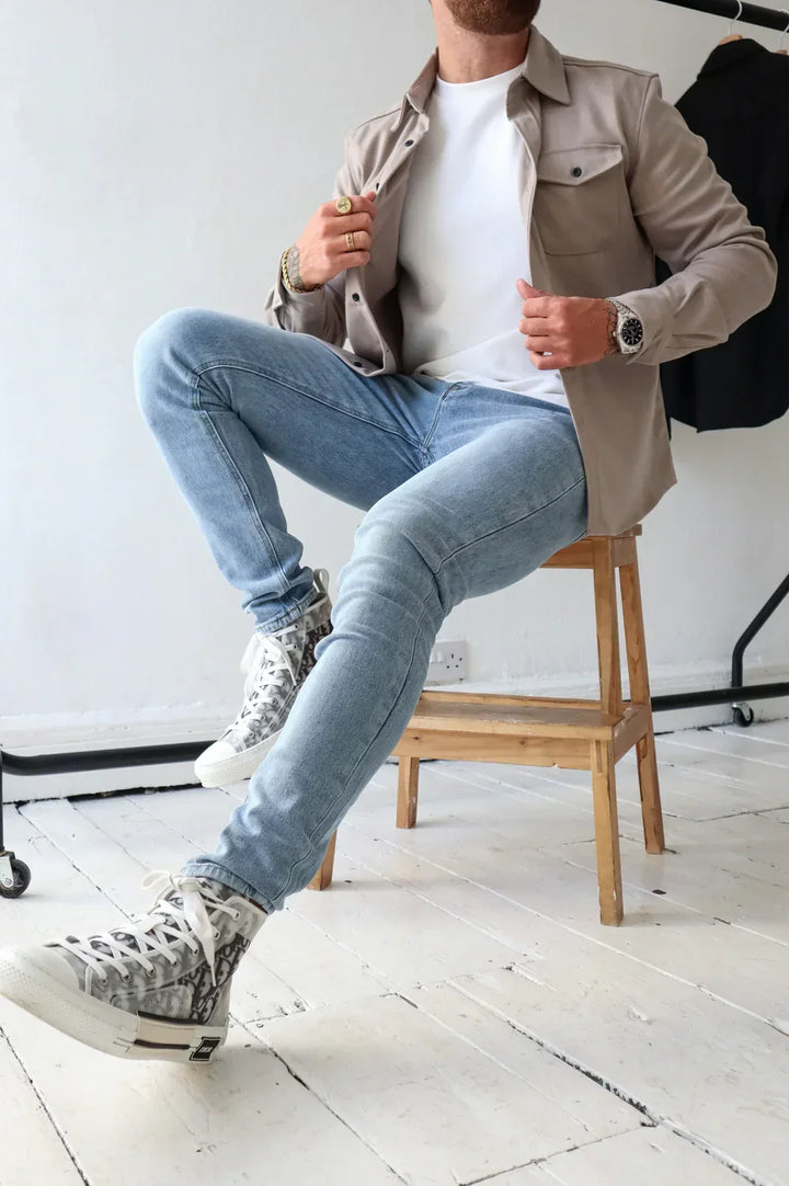 Taupe overshirt with white t-shirt and light jeans, seated on wooden stool indoors.