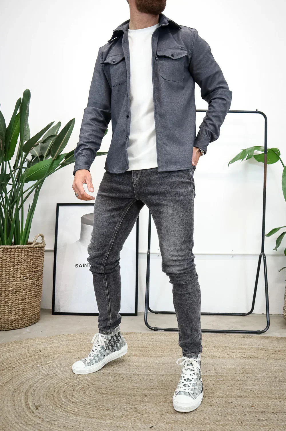 Grey overshirt with white t-shirt and jeans, standing indoors beside plants and art.