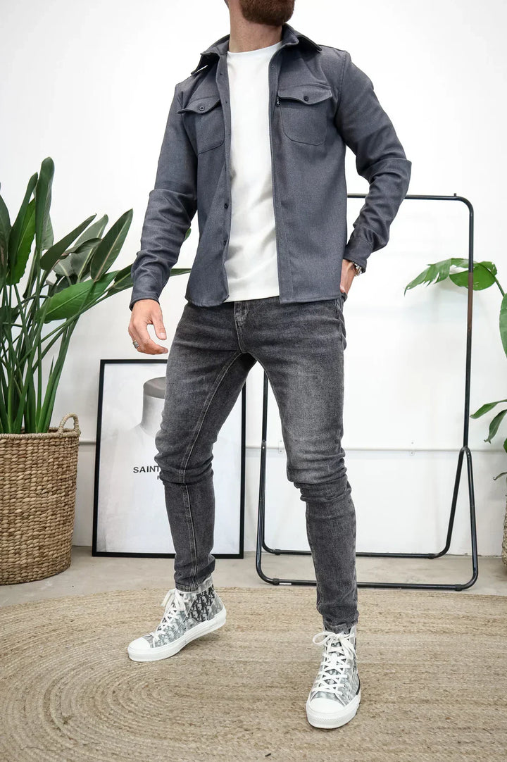 Grey overshirt with white t-shirt and jeans, standing indoors beside plants and art.