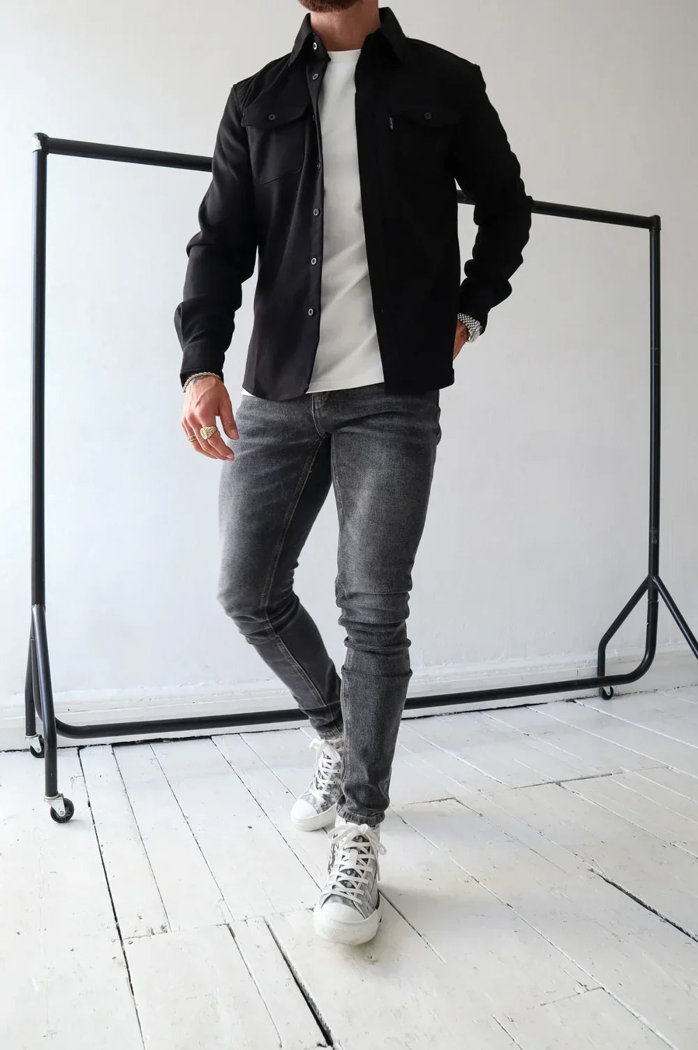 Black overshirt with white t-shirt and jeans, standing in a room with framed art and railing.