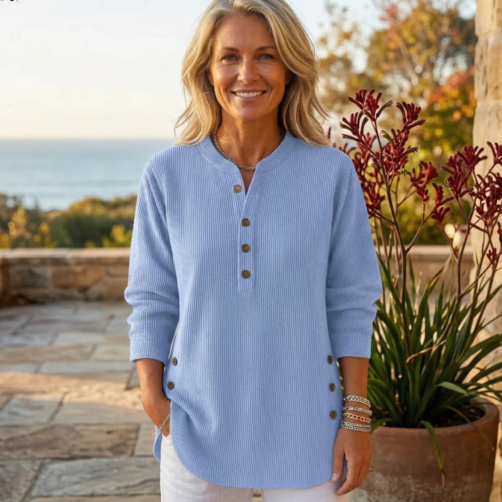Woman wearing a light blue jumper with buttons, in a garden with ocean view and plants.