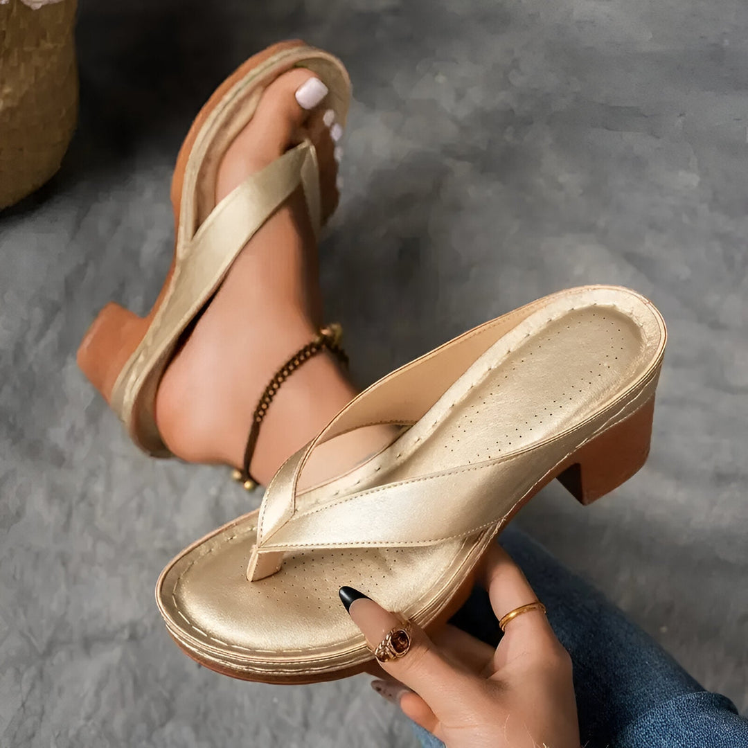 Gold high-heeled mule with perforated insole being held by a hand, worn with anklet on carpet background.