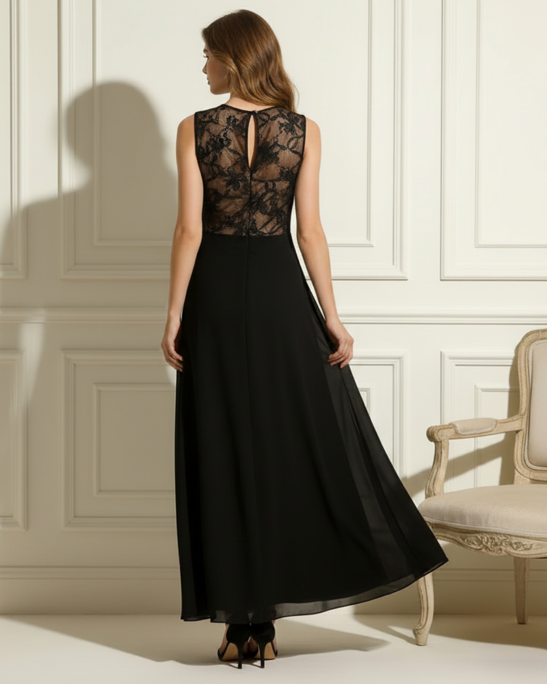Model in black dress with lace back, standing in an elegant room featuring classic paneling and upholstered chair.