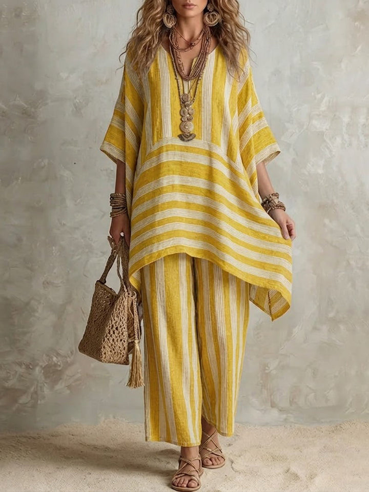 Yellow and white striped two-piece set with flowing top and wide pants, accessorized with layered necklaces and a woven bag, in a neutral studio setting.