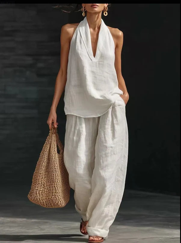 Woman in an elegant white halter top and flowing pants holding a woven bag, standing in a dimly lit urban setting.