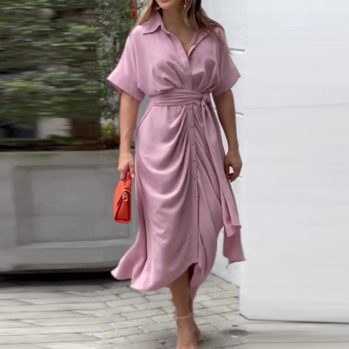 Soft pink wrap maxi dress with subtle drapes, worn while holding a small orange handbag in front of greenery.