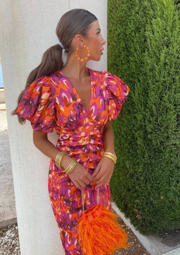 Woman in an orange and purple dress with puff sleeves and feather purse, beside a white column and lush greenery.