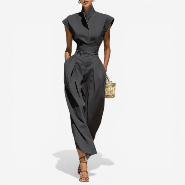 Model in elegant charcoal jumpsuit with high collar and wide waistband, holding a wicker purse against a white background.