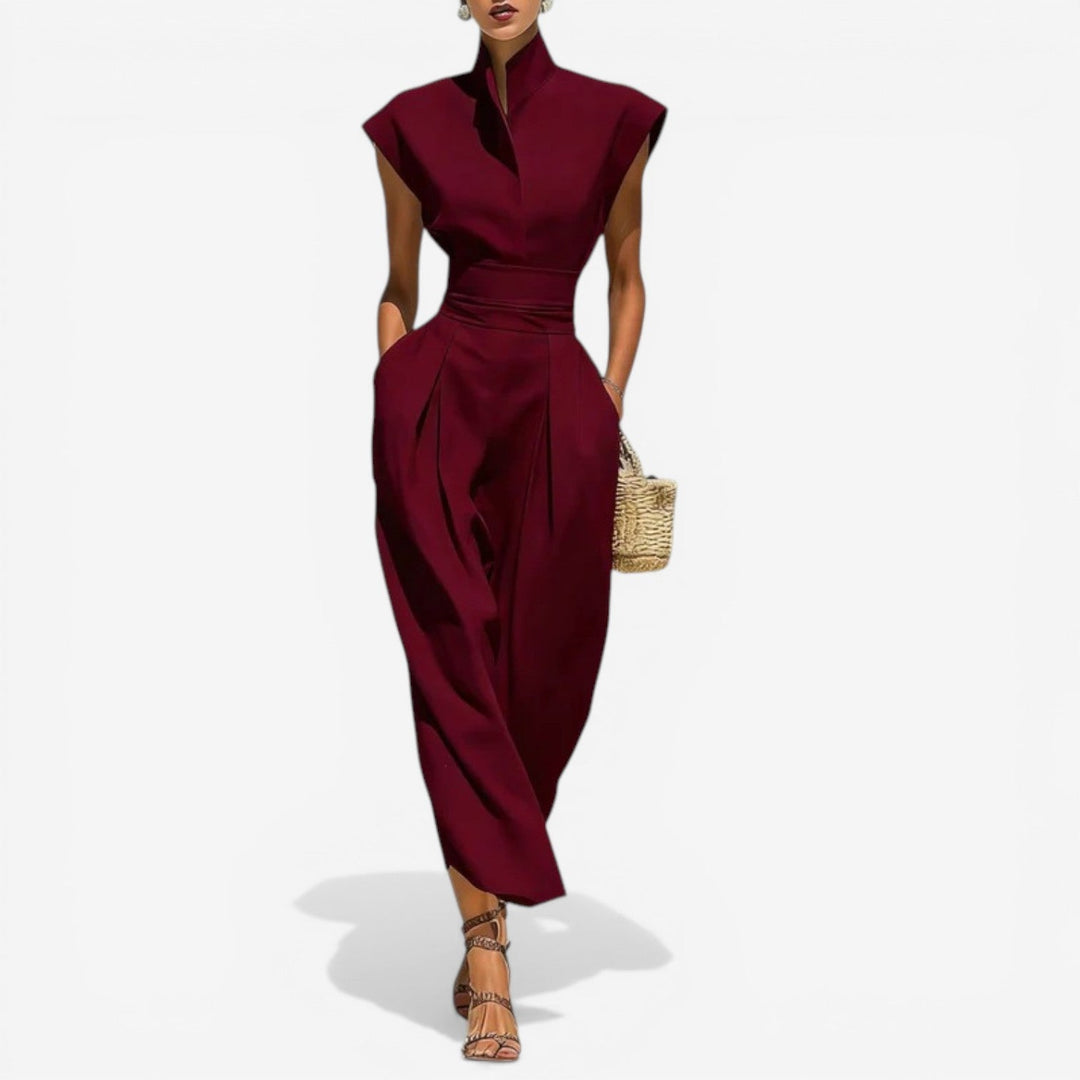 Model in elegant burgundy jumpsuit with high collar and wide waistband, holding a wicker purse against a white background.