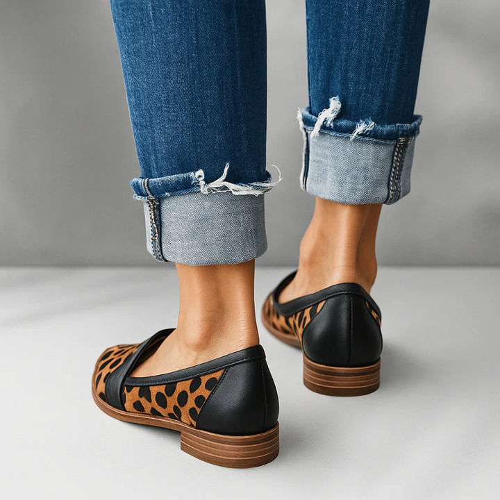 Back view of leopard print orthopedic moccasins with rolled-up dark jeans on a light gray surface.