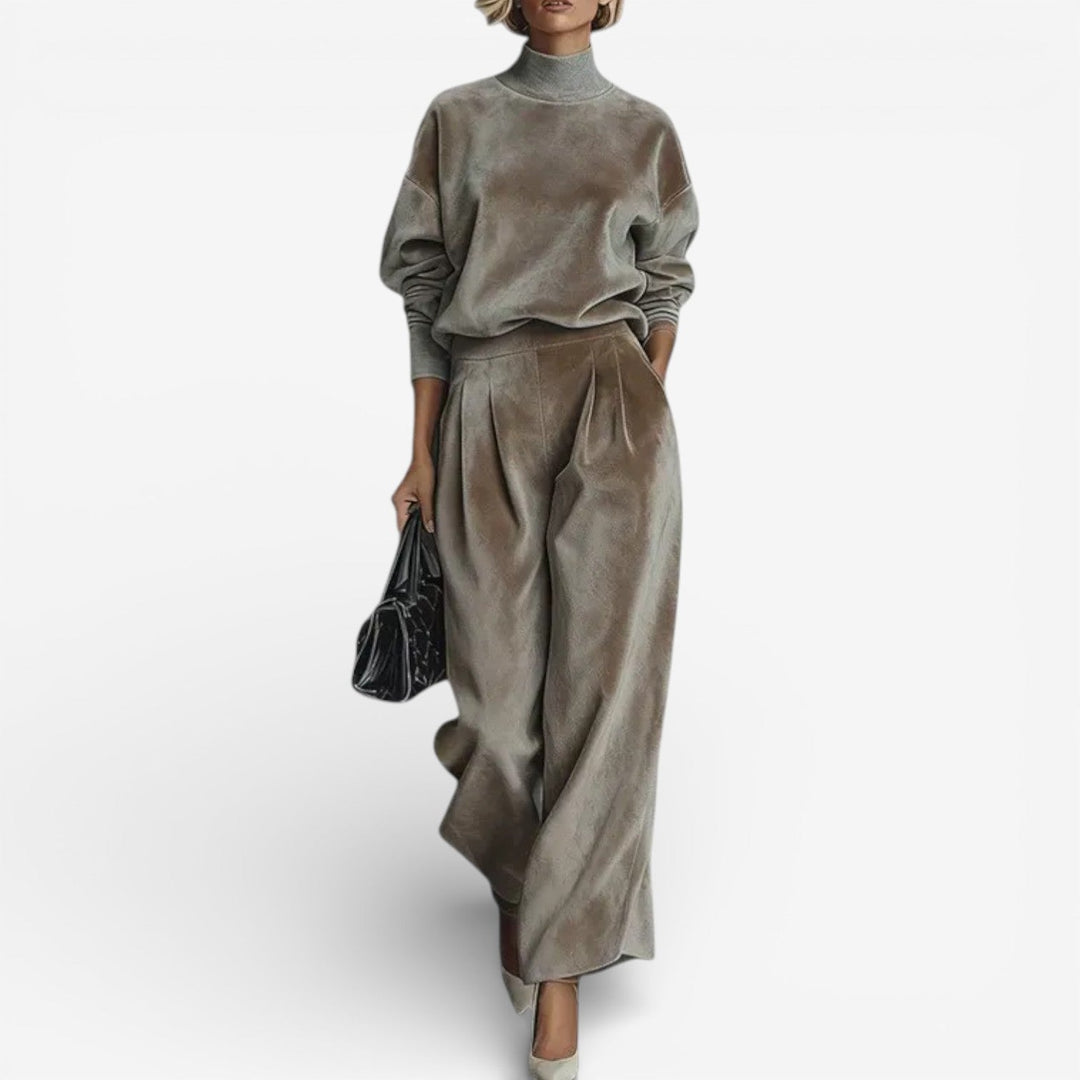 Model in taupe contemporary set with turtleneck and wide-leg pants, holding a black bag, against a white background.